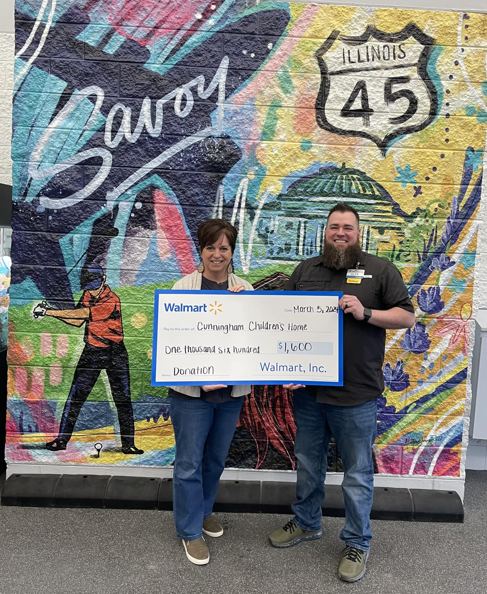 Adult male and female standing in front of a painted wall with illustrations that represent Savoy and holding a large check from Walmart valued at $1,600.