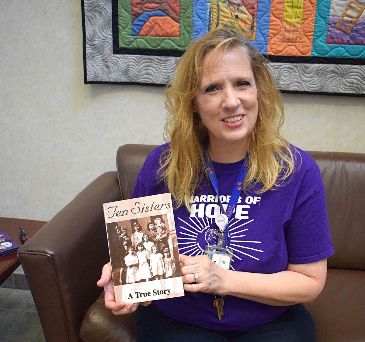 Woman with shoulder length blond hair wearing a purple t-shirt and sitting on a couch proudly displaying a book in her hands