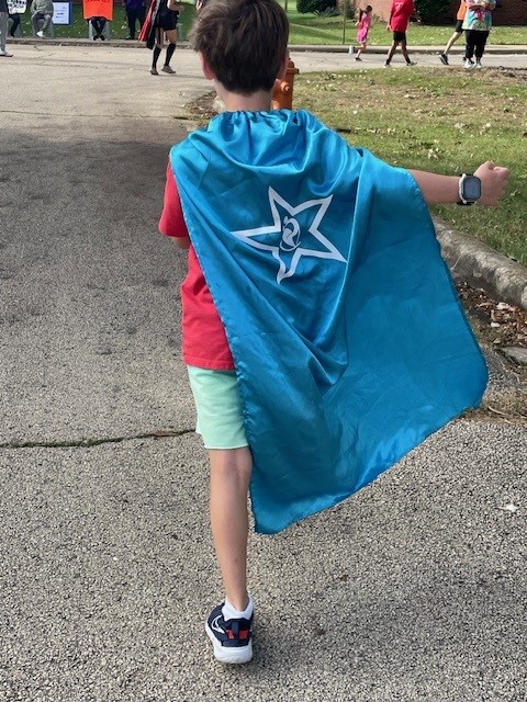 back of a young boy wearing a red t-shirt, light green shorts and an aqua blue superhero cape with a white star and cunningham logo on it