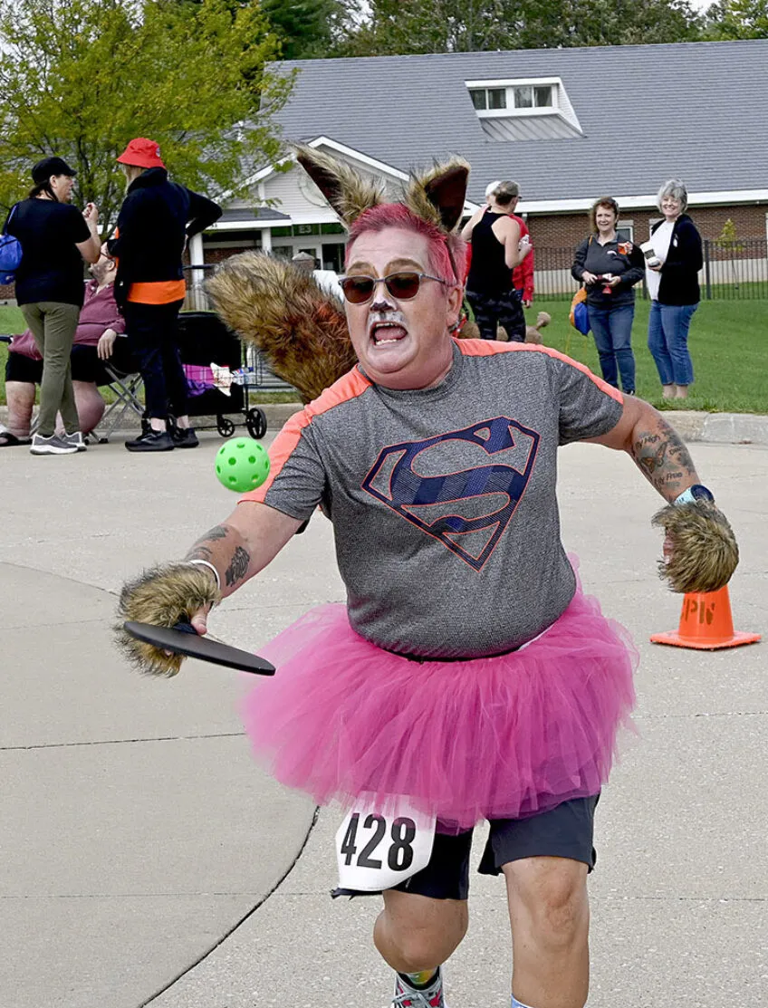 Woman dressed as super squirrel, running while bouncing a ball with a pickle ball paddle.