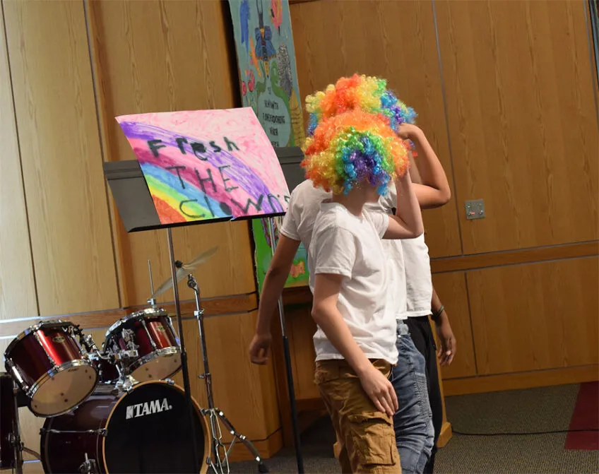 Two young wearing white t-shirts and rainbow color curly wigs standing on a platform performing at a talent show.