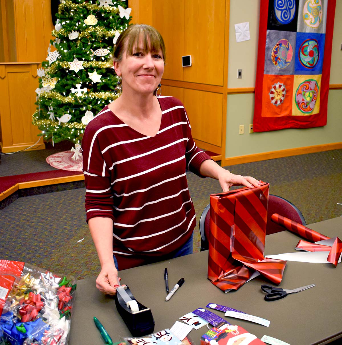 Woman wearing maroon and white striped sweater standing at a table and wrapping a present in the chapel with the Christmas tree behind her in the background.