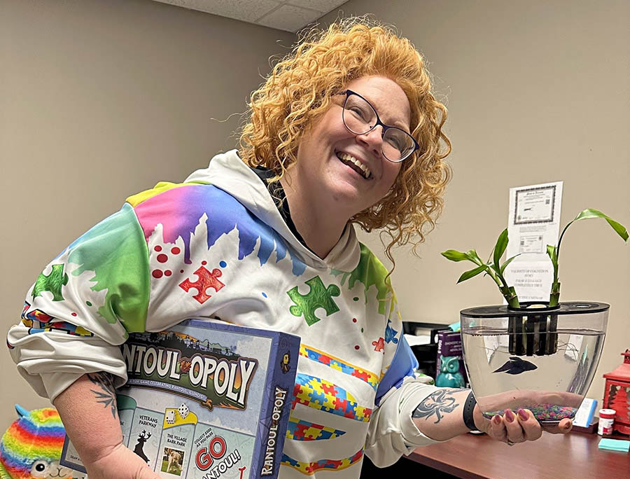 Woman with red curly hair smiling holding a monopoly game and holding a fishbowl with a goldfish in it