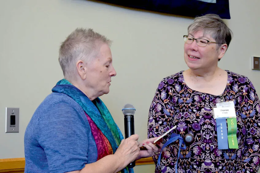 Women wearing blue shirt and colorful scarf reading from a card with a microphone in other hand as another woman in a floral dress listens and smiles intently