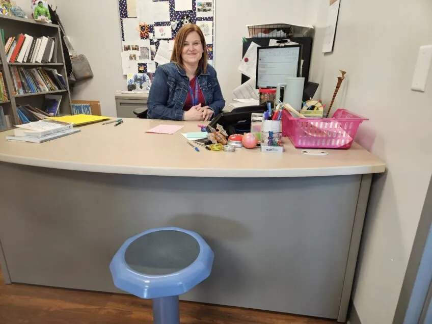 Woman principal sitting at her desk with fidget toys on desk and blue wobble chair in the front of desk