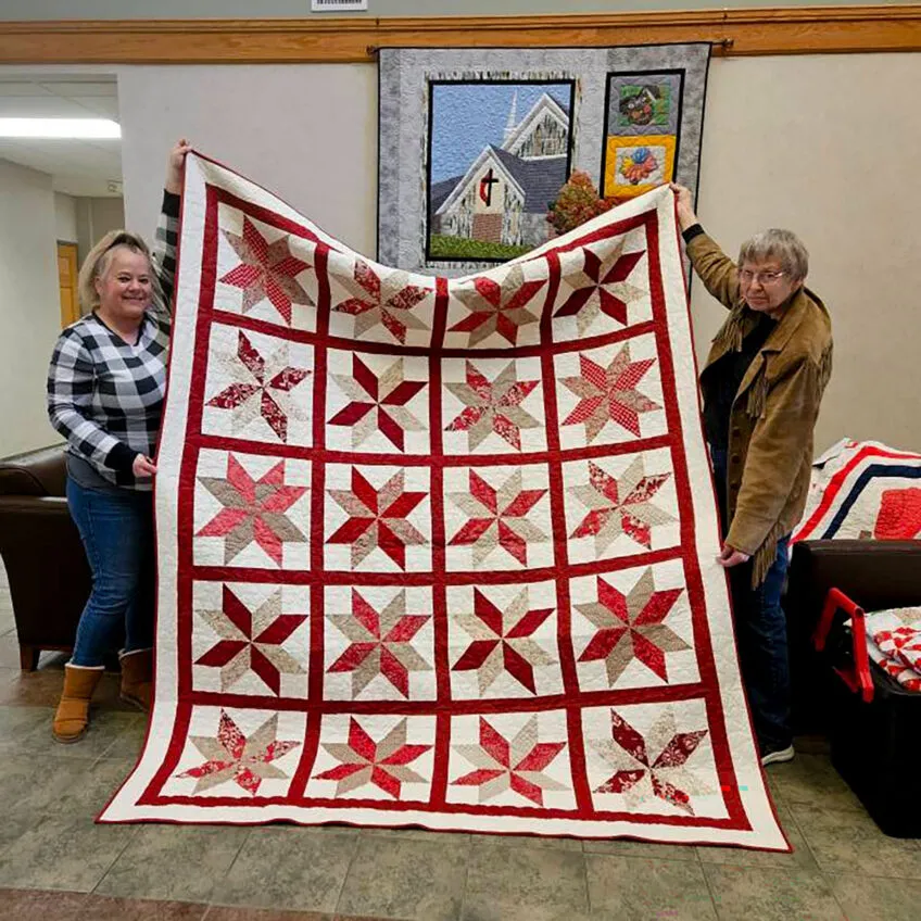 Two women holding up a quilt that has an ivory background and blocks with a star pattern made out of reds and tans
