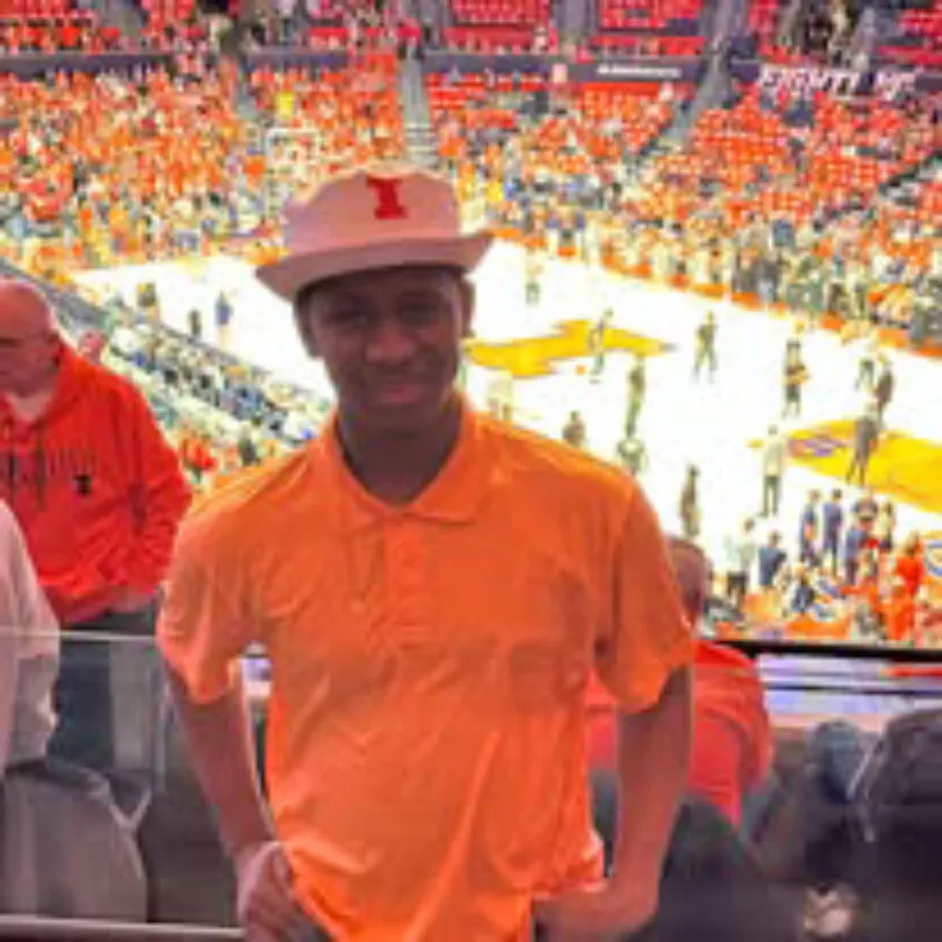 teenage boy decked out in an University of Illinois hat and orange shirt posing with the basketball court and game happening behind him