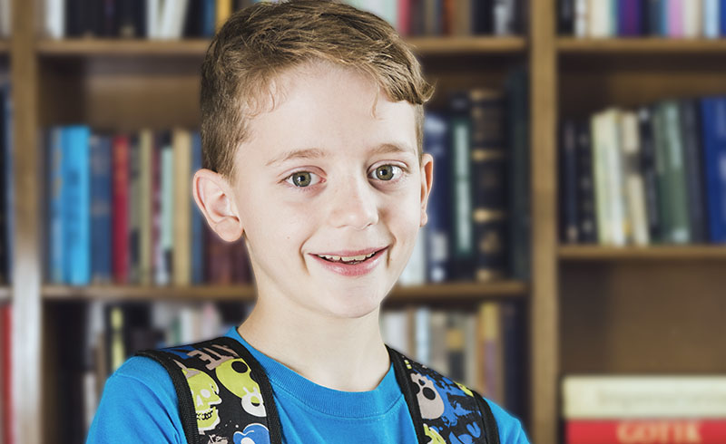 close up of a boy with reddish brown hair, blue shirt and backpack smiling with a bookcase behind him