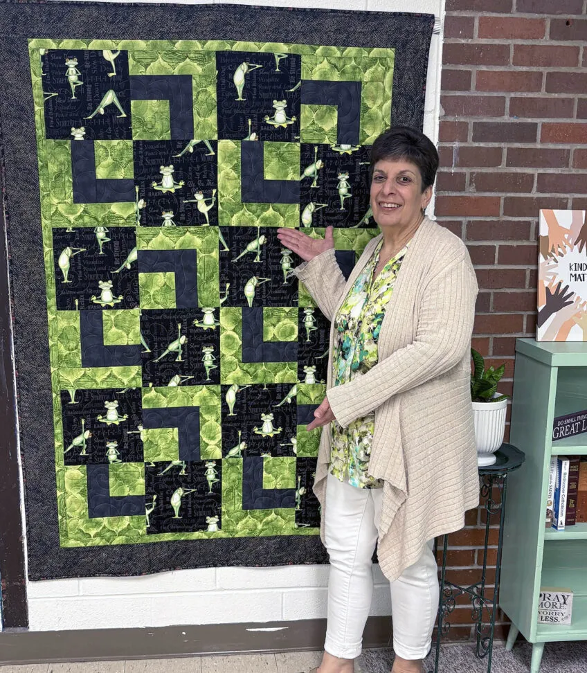 Woman standing in front of a hanging quilt with a green and black pattern background and images of yoga frogs
