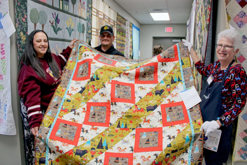 A man and two women holding up a quilt and smiling. Quilt has colors of white, yellow and salmon and pictures of horses