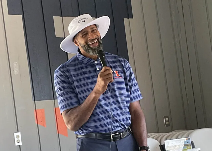 Kendall Gill smiling and talking on a microphone wearing a wide-rimmed sun hat and wearing a blue striped polo