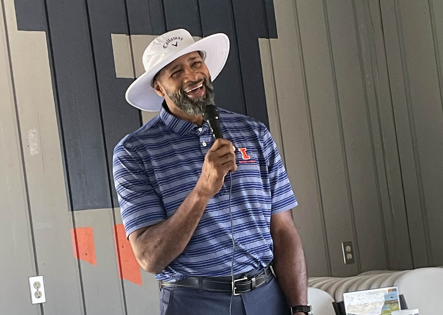 Kendall Gill smiling and talking on a microphone wearing a wide-rimmed sun hat and wearing a blue striped polo