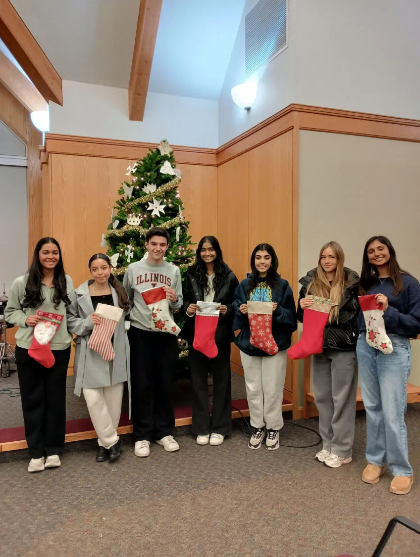 group of college aged young people standing in front of a christmas tree and holding stockings